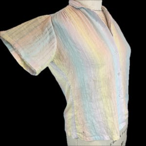 May include: A short-sleeved button-down shirt featuring a vertical striped pattern in soft pastel shades of pink, yellow, blue, and green. The fabric has a subtle crinkled texture, a classic collar, and a row of small white buttons down the front.