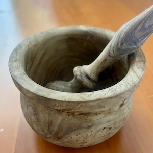 Puede incluir: Un juego de mortero y maja de madera. El mortero es un cuenco redondo con un borde ancho, y la maja es un palo largo y redondeado. La madera tiene un color marrón claro natural con vetas más oscuras.