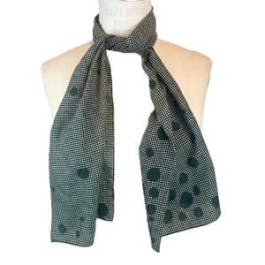 May include: A dark green and white polka dot scarf draped over a mannequin. The scarf has a repeating pattern of small white dots and larger dark green circles. The scarf is made of a lightweight fabric.
