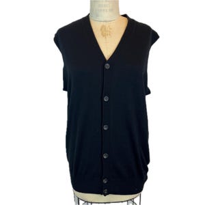 May include: A solid black sleeveless cardigan vest featuring a deep V-neckline and a front button-down closure with five matching black buttons. The garment is constructed with a ribbed hem and arm openings, providing a clean and structured appearance.