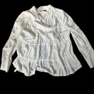 May include: A white long-sleeved blouse featuring a draped cowl neckline and a tiered, ruffled bodice design. The garment is constructed with a lightweight, slightly textured fabric and includes long sleeves with cuffs, creating a layered silhouette.