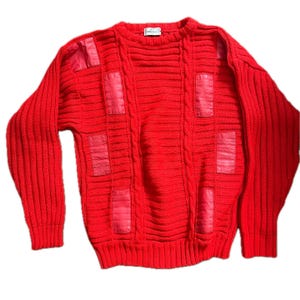 May include: A bright red long-sleeve sweater featuring a textured knit pattern with vertical cable knit columns and smooth rectangular patches applied to the front. The garment has a crew neckline and ribbed cuffs and hem, with a small white label.