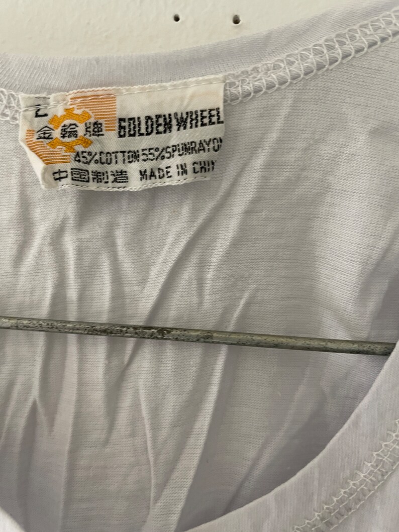 May include: A white t-shirt with a yellow tag that reads "Golden Wheel 45% Cotton 55% Spunrayon Made in China" in English and Chinese.