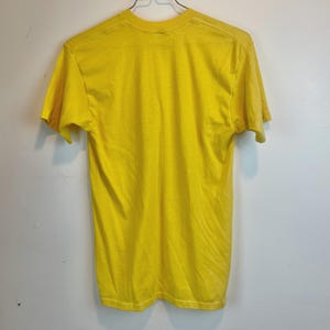 May include: A bright yellow t-shirt with short sleeves, hanging on a metal hanger. The shirt is made of a soft material and has a crew neck. The fabric shows slight wear and tear.