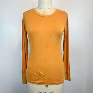Könnte beinhalten: Ein orangefarbenes Langarmshirt mit Rundhalsausschnitt. Das Shirt besteht aus einem weichen, dünnen Material und hat mehrere kleine Löcher. Das Shirt wird auf einer Schaufensterpuppe präsentiert.