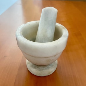 May include: A white marble mortar and pestle set. The mortar is a bowl shape with a rounded base. The pestle is a cylindrical shape. The set is made of a light-colored stone and sits on a wooden surface.