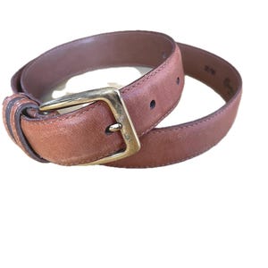 May include: A brown leather belt featuring a rectangular brass-toned metal buckle. The belt strap has a smooth finish with visible stitching along the edges and a series of punched holes for adjustment, coiled into a circular shape.