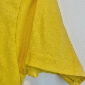 May include: Close-up of a bright yellow t-shirt sleeve. The fabric appears to be a soft knit material. The sleeve is short and has a simple hem. The shirt is a solid color with no visible graphics or patterns.