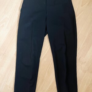 May include: A pair of black trousers with a button closure and pockets. The pants are made of a solid, dark fabric and have a classic, straight-leg design. The trousers are laid flat on a light-colored wooden surface.