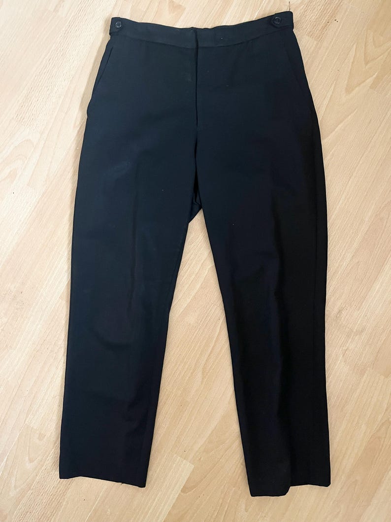 May include: A pair of black trousers with a button closure and pockets. The pants are made of a solid, dark fabric and have a classic, straight-leg design. The trousers are laid flat on a light-colored wooden surface.