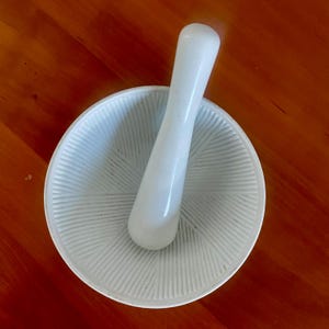 Puede incluir: Un juego de mortero y mano de mortero blancos. El mortero es un cuenco redondo con un interior texturizado, y la mano de mortero tiene una forma cilíndrica larga y lisa. El juego está diseñado para moler y mezclar ingredientes, comúnmente utilizado en la cocina.