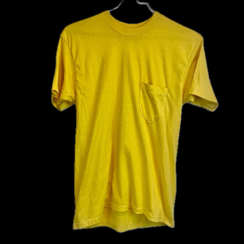 May include: A bright yellow t-shirt with a chest pocket, hanging on a metal hanger against a black background. The short-sleeved shirt has a crew neck and a slightly worn appearance, suggesting it's been washed.