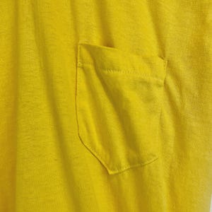 May include: Close-up of a bright yellow t-shirt with a pocket. The pocket is rectangular with rounded corners and is sewn onto the shirt. The fabric has a textured appearance.