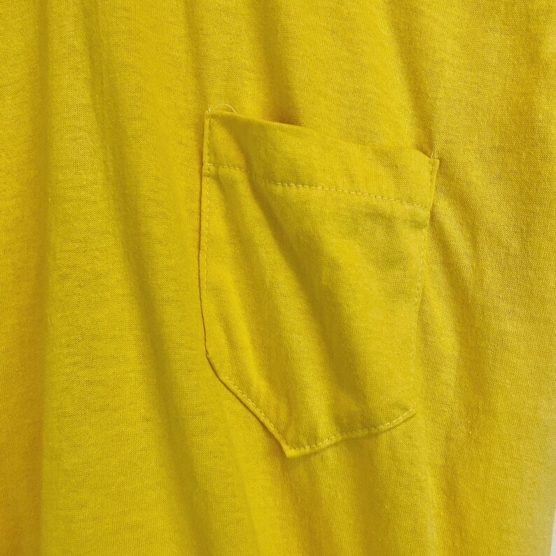 May include: Close-up of a bright yellow t-shirt with a pocket. The pocket is rectangular with rounded corners and is sewn onto the shirt. The fabric has a textured appearance.