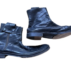 May include: A pair of black leather ankle boots featuring a side zipper closure, a folded collar detail secured with two dark metal snaps, a low stacked heel, and a rounded toe profile. The boots have a smooth finish with visible natural creasing.