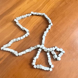 May include: A long strand of small, light gray, cube-shaped beads arranged in a loose, irregular loop. The beads are uniform in size and shape, featuring a matte finish and soft edges, creating a continuous chain that curves across the frame.
