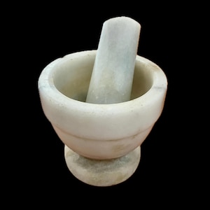 May include: A white marble mortar and pestle set featuring a smooth, rounded bowl with a thick rim and a matching cylindrical pestle. The stone displays subtle grey veining throughout the surface, set against a solid black background.