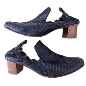 May include: A pair of dark blue, textured leather shoes with a unique design. The shoes have a pointed toe, open sides, and a small, wooden block heel. The leather has a scale-like pattern.