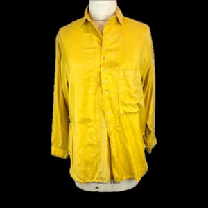 May include: A long-sleeved mustard yellow button-down shirt featuring a pointed collar, a single chest pocket on the left side, and a partial front placket with six visible buttons. The shirt is displayed on a mannequin against a black background.