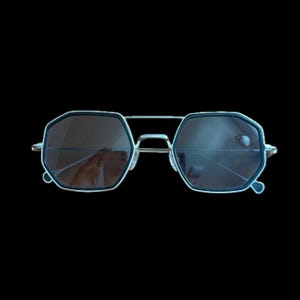 Puede incluir: Gafas de sol geométricas con montura plateada y lentes oscuras. Las lentes son de color marrón oscuro y las monturas tienen una forma octogonal única. Diseño moderno y elegante.