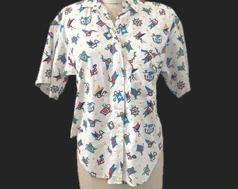 May include: A white short-sleeve button-down shirt featuring a repeating nautical pattern of colorful anchors, ship wheels, and signal flags in shades of red, blue, green, and yellow. The shirt has a classic collar and a single chest pocket.