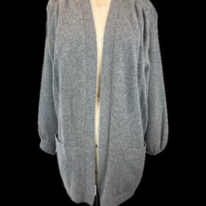 May include: A light gray cardigan sweater with long sleeves and two front pockets. The open-front sweater is displayed on a mannequin against a black background. The sweater has a relaxed fit and a soft, textured appearance.
