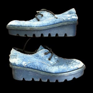 May include: A pair of blue platform shoes with black laces. The shoes have a textured upper and a chunky, ridged sole. The shoes are displayed against a black background.