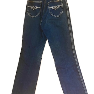 May include: Dark blue denim jeans with decorative stitching and embroidered back pocket designs. The jeans have a classic five-pocket design and a straight leg cut. The back pockets feature an embroidered floral design. The jeans are a vintage style.
