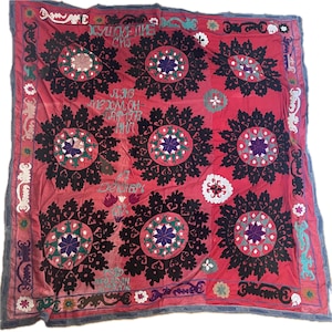 May include: A rectangular textile wall hanging featuring a vibrant red background with large, repeating black circular floral motifs. The design includes intricate embroidery in purple, white, and green, along with Cyrillic text and decorative borders.