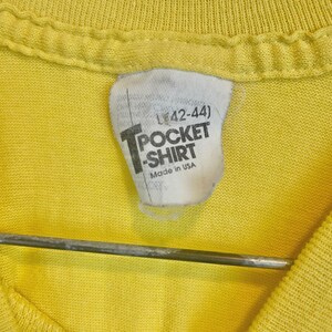 May include: Close-up of a yellow T-shirt with a white tag. The tag reads "L (42-44)" above the words "POCKET SHIRT" and "Made in USA". The shirt is hanging on a metal hanger.