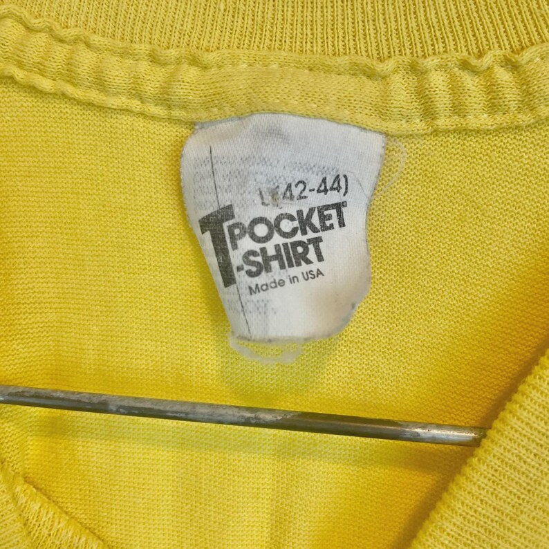 May include: Close-up of a yellow T-shirt with a white tag. The tag reads "L (42-44)" above the words "POCKET SHIRT" and "Made in USA". The shirt is hanging on a metal hanger.