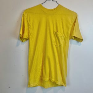 May include: A bright yellow t-shirt with a chest pocket. The short-sleeved shirt is hanging on a metal hanger against a white wall. The shirt is made of a soft material.