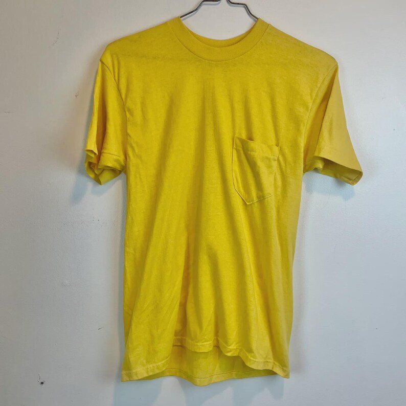 May include: A bright yellow t-shirt with a chest pocket. The short-sleeved shirt is hanging on a metal hanger against a white wall. The shirt is made of a soft material.