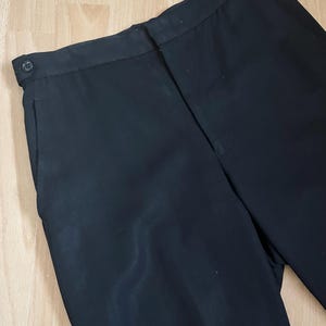 May include: Black dress pants with a button closure and side pockets. The pants are made of a smooth, dark fabric. The image shows a close-up of the waist and upper leg area of the pants.