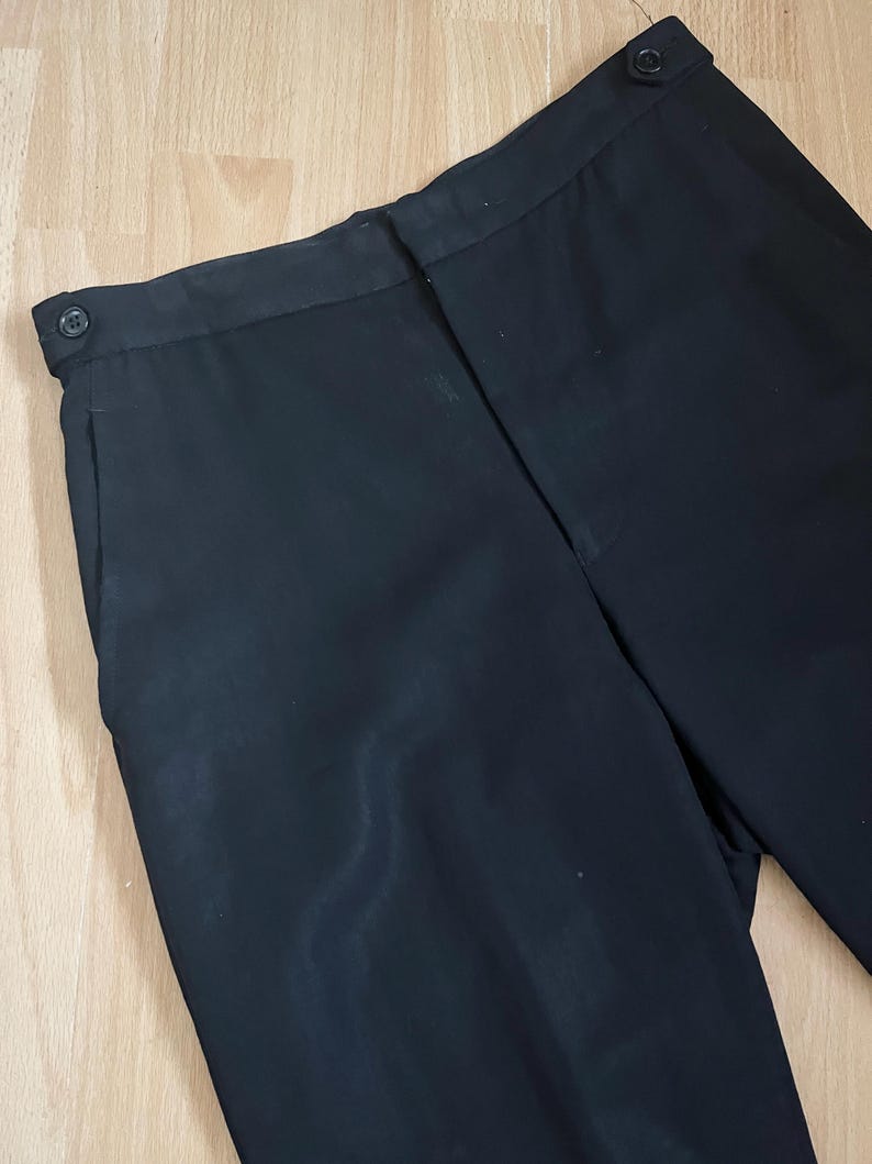 May include: Black dress pants with a button closure and side pockets. The pants are made of a smooth, dark fabric. The image shows a close-up of the waist and upper leg area of the pants.