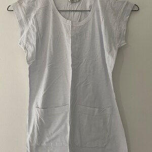 May include: A light gray t-shirt with short sleeves and two patch pockets. The shirt has a round neckline and is hanging on a hanger.