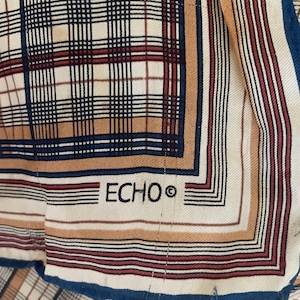 May include: A white silk scarf with a brown, red, and blue plaid pattern. The scarf has a navy blue border and the word "ECHO" printed in black in the center.