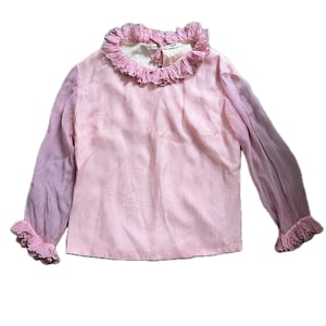 May include: A vintage pink blouse with long sleeves and a ruffled collar and cuffs. The sleeves are a lighter shade of pink than the body of the blouse. The blouse has a delicate, feminine design.