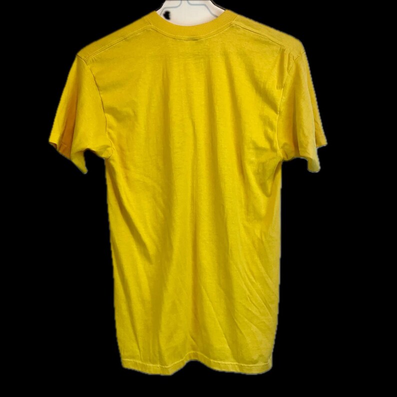 May include: A solid yellow t-shirt, hanging on a white hanger against a black background. The shirt is made of a soft material and has short sleeves and a crew neck. The back of the shirt is visible.