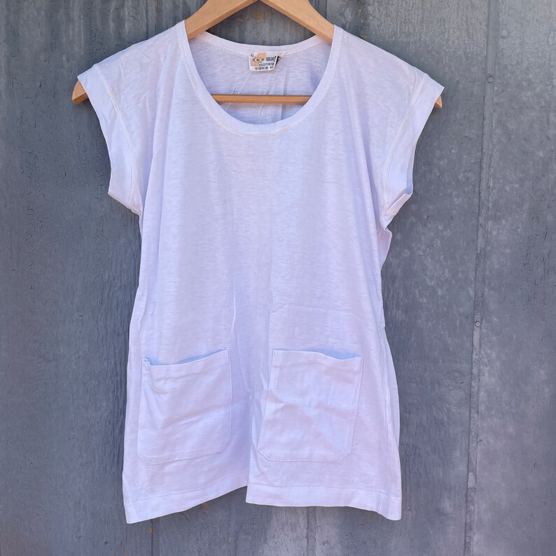 May include: White cotton t-shirt with short sleeves and two patch pockets. The shirt has a round neckline and a relaxed fit.
