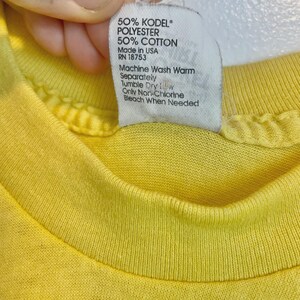 May include: Close-up of a yellow t-shirt with a white tag. The tag lists the fabric composition as 50% KODEL polyester and 50% cotton. It also includes washing instructions: machine wash warm, tumble dry low, and only non-chlorine bleach when needed.