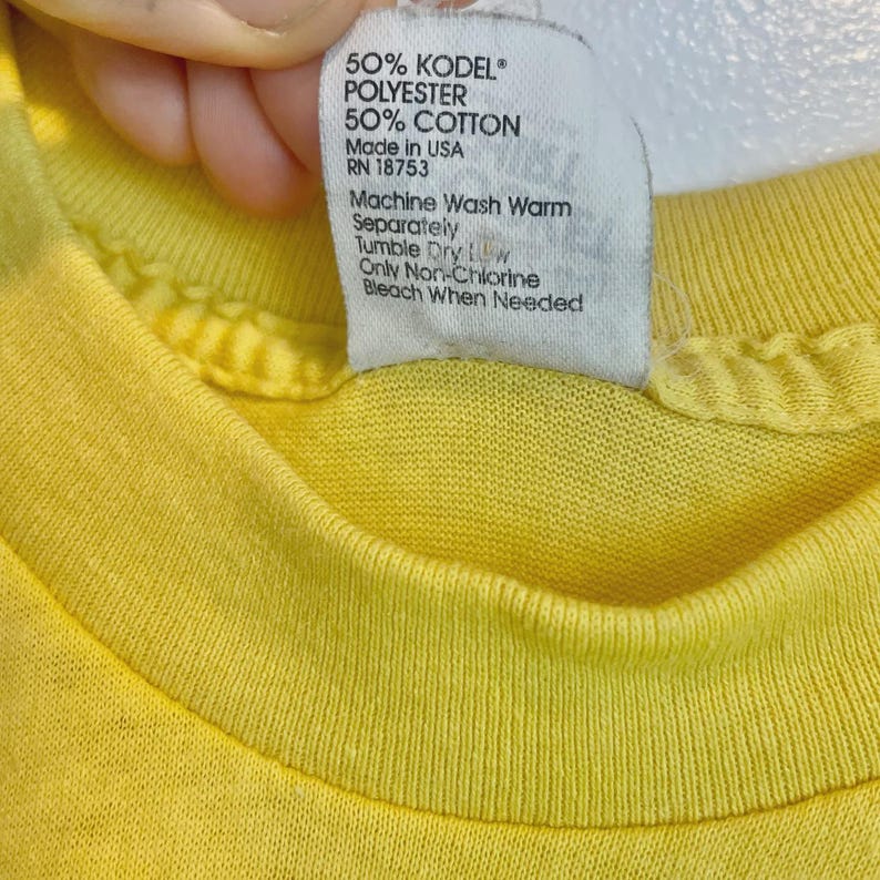 May include: Close-up of a yellow t-shirt with a white tag. The tag lists the fabric composition as 50% KODEL polyester and 50% cotton. It also includes washing instructions: machine wash warm, tumble dry low, and only non-chlorine bleach when needed.