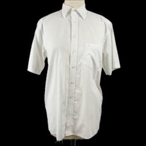 Puede incluir: Camisa blanca de manga corta con cuello y bolsillo en el pecho, expuesta sobre un maniquí. La camisa tiene una botonadura y está colocada sobre un fondo negro. La camisa tiene un estilo clásico e informal.