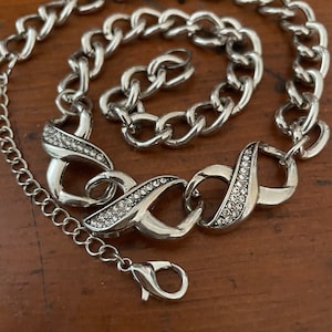 May include: A silver chain necklace with a decorative pendant featuring a twisted design and clear rhinestones.