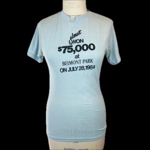 May include: Pale blue t-shirt with short sleeves. The shirt displays the text "almost I WON £75,000 at BELMONT PARK ON JULY 28, 1984" in black lettering. The t-shirt is shown on a mannequin.