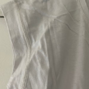 May include: White sleeveless shirt with a stitched seam detail around the neckline and armholes.