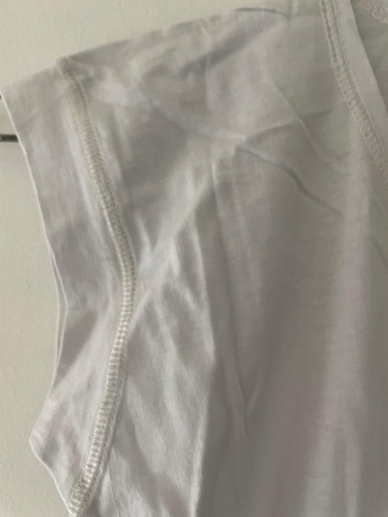 May include: White sleeveless shirt with a stitched seam detail around the neckline and armholes.