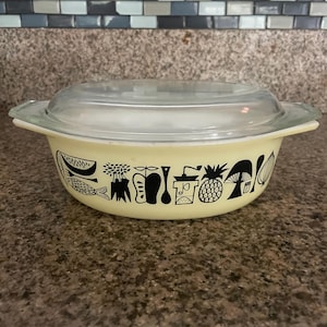 May include: A yellow casserole dish with a black and white design of various fruits, vegetables, and kitchen utensils. The dish has a glass lid.