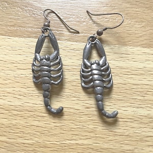 May include: A pair of silver-toned metal earrings featuring a detailed scorpion charm design. Each earring is suspended from a simple curved ear wire hook. The scorpion charms have a textured finish with visible segments, pincers, and a curved tail.