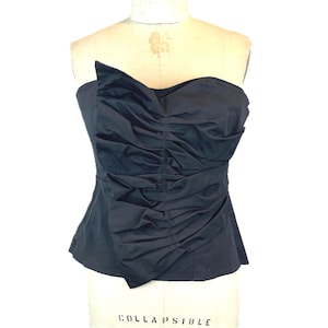May include: A strapless black top featuring a fitted silhouette and a prominent vertical panel of gathered fabric down the center front. The garment is displayed on a dress form, showcasing the textured, ruched detailing across the bodice.